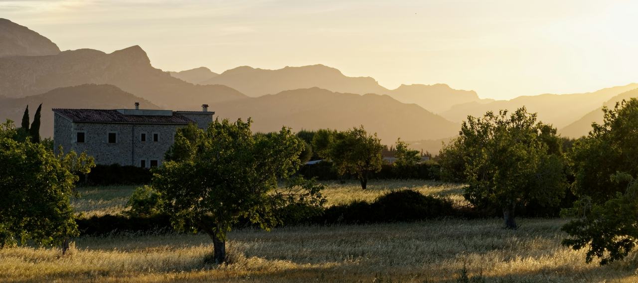 Casa de piedra entre árboles y campo, con la sierra al fondo